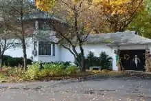 A small white brick gatehouse in front a forest of changing leaves, with a black asphalt driveway on its left