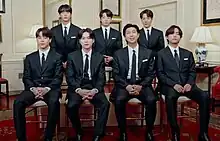 A group of seven South Korean men wearing suit and tie pose for the camera while sitting on their chairs