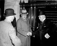 Two older white men in suits, hats, and eyeglasses shake hands. The one on the left looks towards the foreground, where a third white man in a hat looks back, his back to the camera.