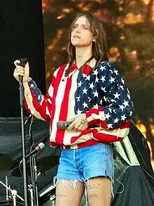 A woman wears an American flag shirt and short jean shorts as she holds a microphone and looks into the distance.