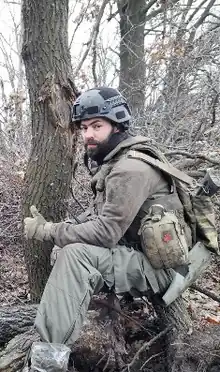 Mangushev in Olive green military clothing and a ballistic helmet, with a rifle over his shoulder and a first aid kit on his hip giving a thumbs-up signal towards the photographer