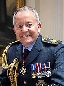 Portrait photograph of head and chest of Sir Richard Knighton, taken indoors in front of a display banner.  Knighton is wearing his Royal Air Force 'Number 1' service dress without head-dress, with medal ribbons, but without actual medals, and above the ribbons is his name badge.  Knighton has a broad smile, clean-shaven, and brushed-back light-coloured hair.