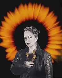 Bust-length hand-coloured photocollage portrait by Ming de Nasty of Rhonda Wilson in a leather jacket with a halo formed by a sunflower and holding proteas