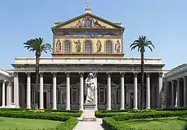 A courtyard with palm trees and a greater-than-lifesized statue of Saint Paul holding a sword in front of the colossal portico of the basilica and a large mural covering the upper facade