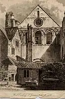 Monochrome illustration of the west end of St James' church, published in 1820. The image shows the parts of the priory stonemasonry incorporated into the church facade and a jumble of non-ecclesiastical buildings in the foreground including a tall chimney.