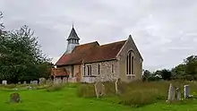 A small stone church with red tiled roofs seen through a churchyard from the south-east. On its far gable is a bellcote with a pyramidal roof