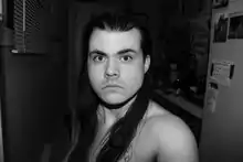 Black and white headshot of B. G-Osborne, a white person with long hair and an intense stare. Landscape picture orientation with head placed slightly up from centre of image. Body takes up middle third of picture plane. They are not smiling and looks directly out at the viewer with their left shoulder forward and in the foreground. Artist has long hair, worn slicked back on top, tucked behind their ears, allowing the hair to fall over chest. There is a shadow of stubble and facial hair. To the left is a set of blinds and to the right a wall with taped notes and photos that gives way to a counter